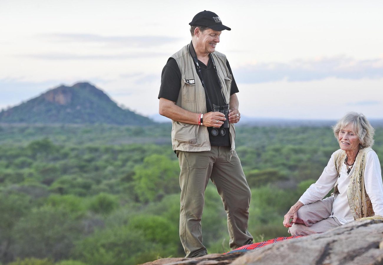 Will Travers stood next to Virginia McKenna, who is sitting down on a rock with a mountainous Kenyan landscape in the background